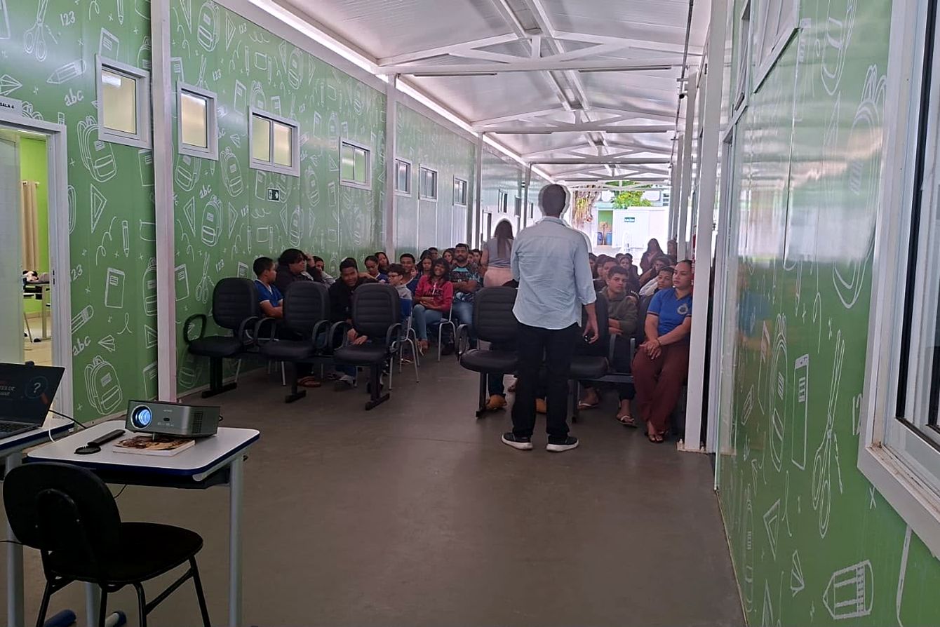 Palestrante de costas, diante da plateia de estudantes, com projetor em primeiro plano